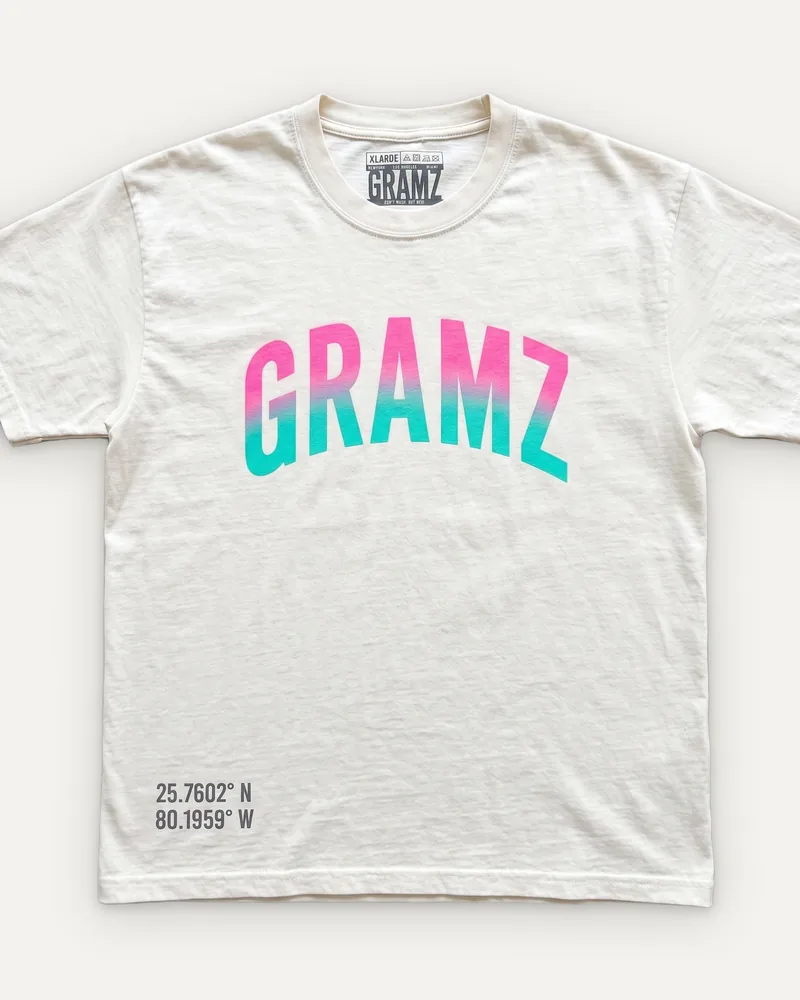 Design rendering: cream tee with an arched GRAMZ chest print fading from pink to teal and Miami coordinates at the hem